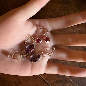May include: Three pairs of vintage clip-on earrings with red and purple stones. The earrings are made of gold-toned metal.