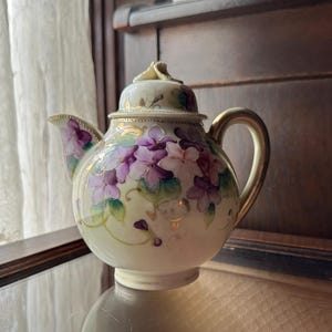 May include: A small, cream-coloured teapot with a floral design. The teapot is decorated with purple and pink flowers and gold accents. The teapot has a lid and a handle.