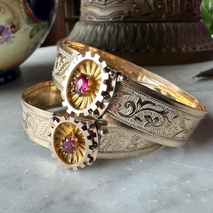 Peut inclure: Deux bracelets rigides en métal doré ornés de gravures florales complexes sur toute la bande. Chaque bracelet est centré par un ornement circulaire en forme d'engrenage avec un motif rayonnant et une pierre précieuse rose foncé ovale.