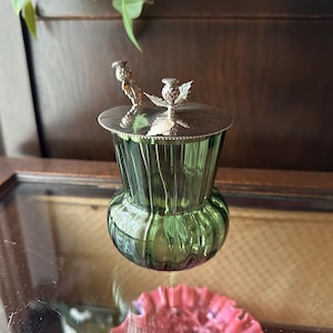Peut inclure: Un pot en verre vert avec un couvercle argenté et des figurines d'oiseaux décoratives. Le pot a un design côtelé et une base arrondie. Le couvercle est argenté avec un bord festonné. Les figurines ajoutent de l'élégance.