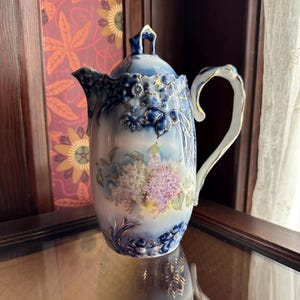 May include: A vintage porcelain coffee pot with a blue and white floral design. The pot has a tall, slender shape, a curved handle, and a lid with a decorative finial. The floral pattern includes purple and white flowers with gold accents.