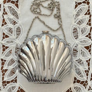 Peut inclure: Un sac pochette rigide en forme de coquillage de couleur argentée avec une finition métallique et des détails nervurés verticaux. Le sac présente une fermeture supérieure à charnière et une fine bandoulière en chaîne métallique assortie.