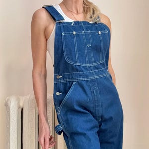 May include: Dark blue denim overalls with white stitching. The overalls have adjustable straps, multiple pockets, and metal button closures. The garment is a classic workwear style.
