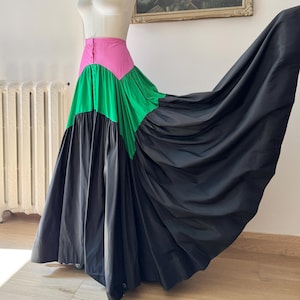 May include: A floor-length skirt featuring a high-waisted pink band, a vibrant green middle section, and a voluminous black lower panel. The skirt is displayed on a mannequin, showcasing a full, gathered silhouette with a smooth, solid finish.