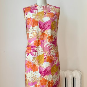 May include: A sleeveless shift dress featuring a vibrant floral and tropical leaf pattern in shades of bright pink, orange, and golden yellow on a white background. The dress has a high neckline, a straight silhouette, and two horizontal slit pockets.