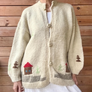 May include: A cream-coloured knit cardigan featuring a button-front closure with three round knit buttons. The garment displays a landscape pattern along the bottom hem, including brown houses with red roofs, small green trees, and brown pine trees.