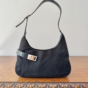 May include: A black hobo-style shoulder bag crafted from a durable fabric material. The bag features a single adjustable strap and a prominent silver-toned metal buckle closure on the front, which includes a small engraved logo detail.