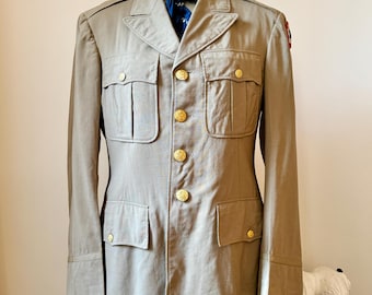 Op de afbeelding: Een beige militaire jas met gouden knopen en een patch op de rechtermouw. De jas heeft vier zakken en een kraag.