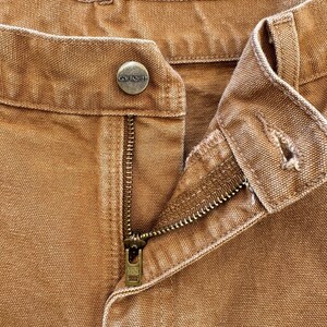 May include: Close-up of brown work pants with a button closure and a zipper. The button is embossed with the brand name "Carhartt". The zipper is partially open, revealing the brass-colored metal teeth. The fabric is a sturdy, textured material.