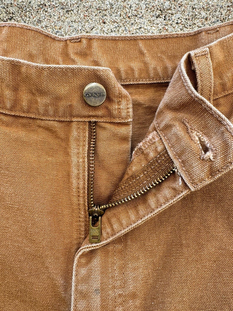 May include: Close-up of brown work pants with a button closure and a zipper. The button is embossed with the brand name "Carhartt". The zipper is partially open, revealing the brass-colored metal teeth. The fabric is a sturdy, textured material.