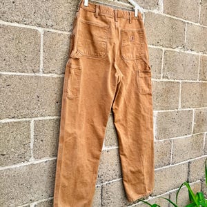 May include: Brown work pants with multiple pockets, hanging on a white hanger against a brick wall. The pants have a rectangular logo on the back pocket. The pants are made of a durable material.