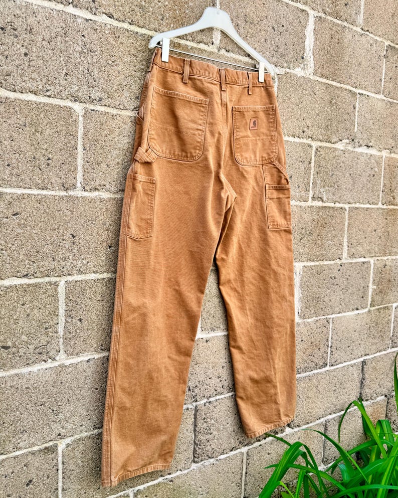 May include: Brown work pants with multiple pockets, hanging on a white hanger against a brick wall. The pants have a rectangular logo on the back pocket. The pants are made of a durable material.
