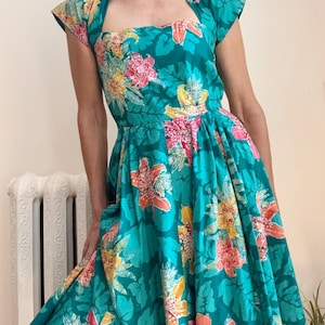 May include: A teal dress featuring a vibrant floral print with pink, orange, and yellow flowers and green foliage. The dress has a square neckline, cap sleeves, and a full, pleated skirt that falls below the knee, creating a structured silhouette.