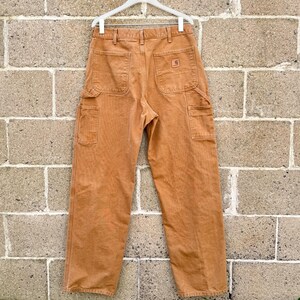 May include: A pair of tan work pants with multiple pockets, hanging on a white hanger against a brick wall. The pants have a classic workwear design, featuring a logo patch on the back pocket.