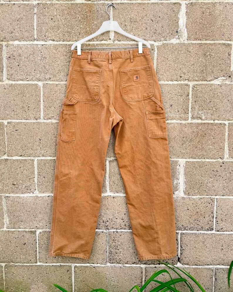 May include: A pair of tan work pants with multiple pockets, hanging on a white hanger against a brick wall. The pants have a classic workwear design, featuring a logo patch on the back pocket.
