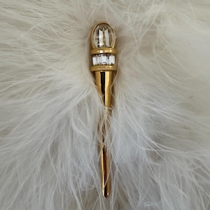 May include: A gold-toned brooch with a torch design. The top features a large, clear, oval-shaped crystal above a row of smaller rectangular crystals. The brooch is set against a backdrop of white feathers.