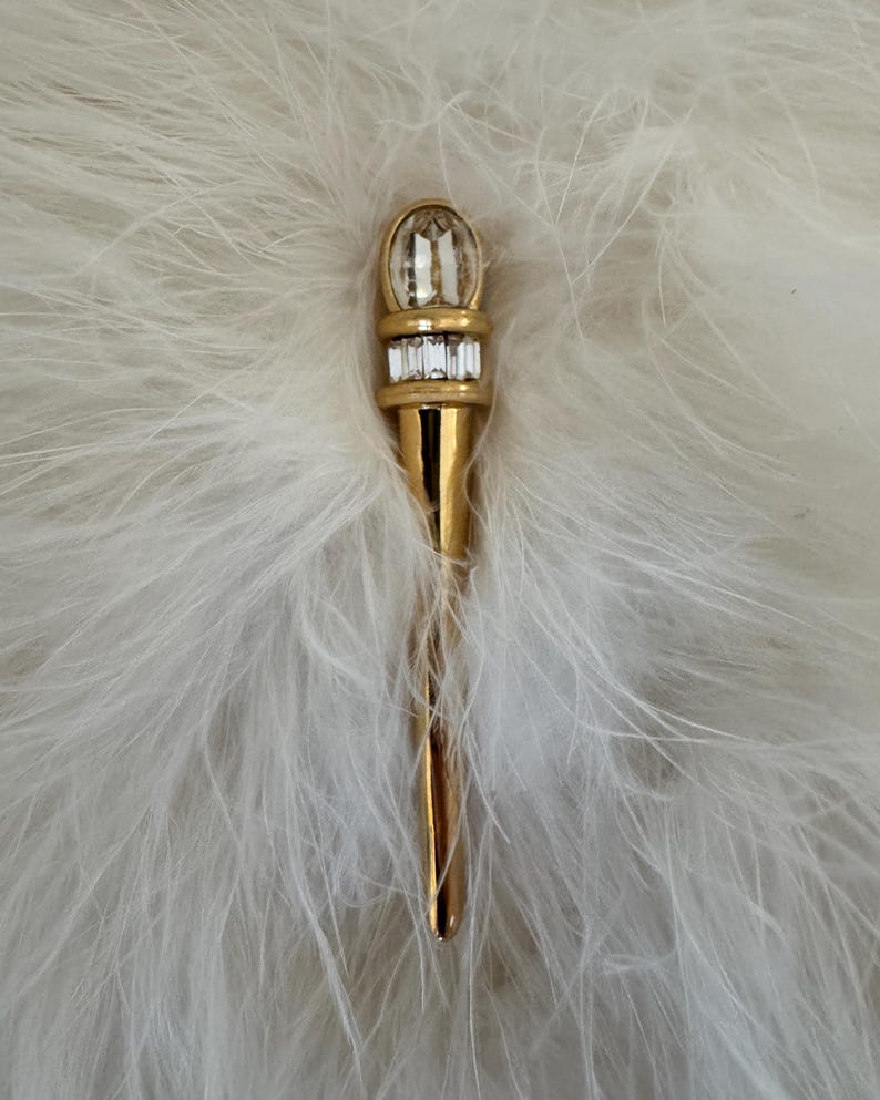 May include: A gold-toned brooch with a torch design. The top features a large, clear, oval-shaped crystal above a row of smaller rectangular crystals. The brooch is set against a backdrop of white feathers.
