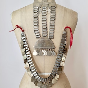 May include: A silver-toned metal necklace featuring three vertical rows of rectangular links connected to a central plate with dangling circular charms. A long chain of rectangular links with round engraved pendants hangs below, accented by red yarn ties.
