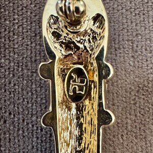 May include: Close-up of a gold-toned brooch with an elongated, textured design. The brooch features a decorative top and a central oval with the initials "RR" in black. The overall design is ornate and detailed.
