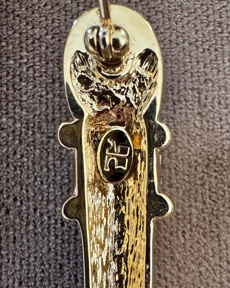 May include: Close-up of a gold-toned brooch with an elongated, textured design. The brooch features a decorative top and a central oval with the initials "RR" in black. The overall design is ornate and detailed.