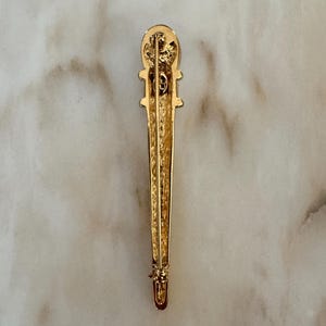 May include: A gold-toned brooch with an elongated, vertical design. The brooch features a detailed top section with a decorative element and a pin closure at the bottom. The surface has a textured appearance, and the overall style is vintage.