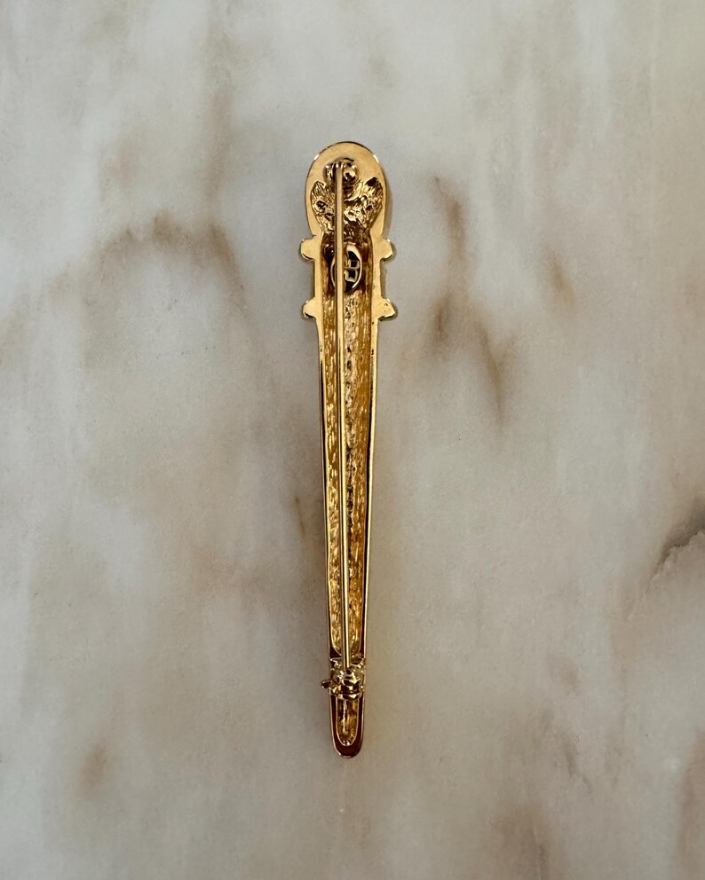 May include: A gold-toned brooch with an elongated, vertical design. The brooch features a detailed top section with a decorative element and a pin closure at the bottom. The surface has a textured appearance, and the overall style is vintage.