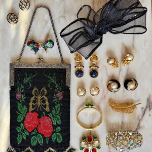 May include: An assortment of vintage jewelry and accessories, including a beaded black purse with red roses, various earrings, a black mesh bow, a gold bracelet, and a jeweled brooch. The items are arranged on a marble surface.
