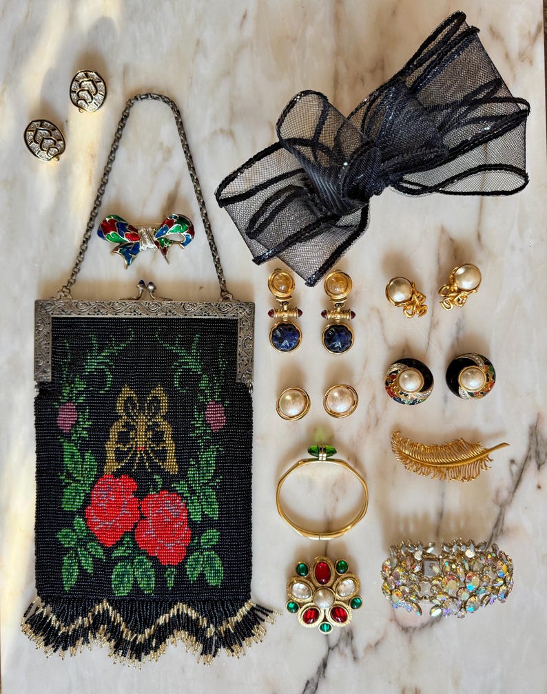 May include: An assortment of vintage jewelry and accessories, including a beaded black purse with red roses, various earrings, a black mesh bow, a gold bracelet, and a jeweled brooch. The items are arranged on a marble surface.