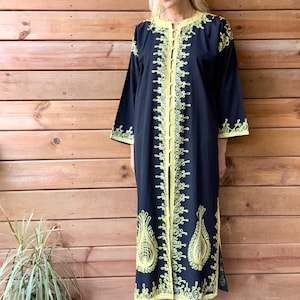 May include: A long-sleeved black kaftan featuring a round neckline and a central vertical row of buttons. The garment is decorated with intricate yellow embroidery patterns along the front placket, cuffs, and hemline, with side slits.