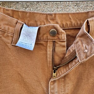 May include: Close-up of Carhartt brown work pants. The pants have a button closure, a zipper, and a label with the Carhartt logo and size information. The pants are made of a durable, heavy-duty fabric.