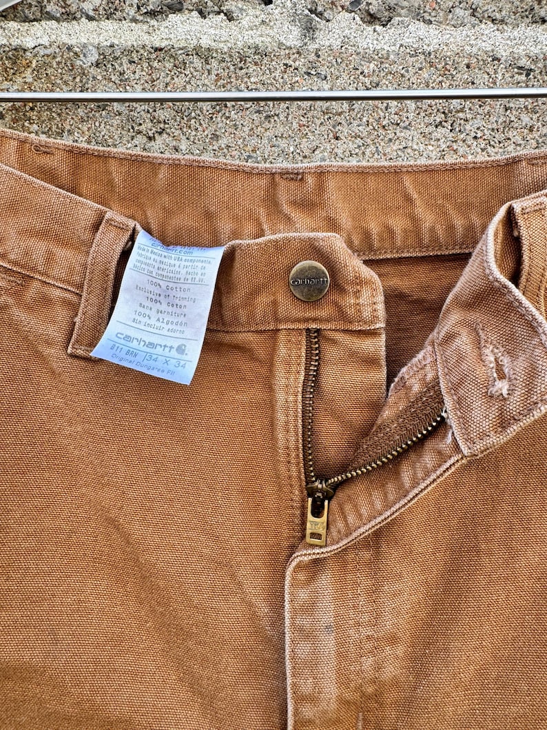 May include: Close-up of Carhartt brown work pants. The pants have a button closure, a zipper, and a label with the Carhartt logo and size information. The pants are made of a durable, heavy-duty fabric.