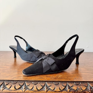 Peut inclure: Une paire d'escarpins noirs à bride arrière avec petits talons et bouts carrés. Chaque chaussure présente un nœud sur le cou-de-pied. Les chaussures sont présentées sur une surface en bois.