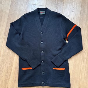 May include: A black cardigan sweater with a V-neck, button closure, and two front pockets. The sweater has orange stripes on the sleeves and orange trim on the pockets. The sweater is made of a textured knit fabric.