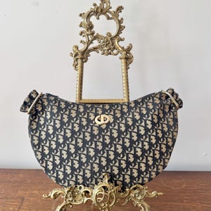May include: A navy blue and beige handbag with a repeating pattern, displayed on a decorative gold-colored stand. The bag has a curved shape and a gold-colored clasp. The stand is ornate with scrollwork.