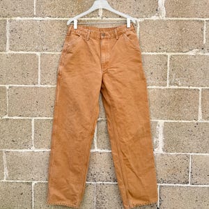 May include: A pair of tan-colored work pants hanging on a white hanger against a brick wall. The pants have a classic five-pocket design with reinforced stitching and a straight leg. The fabric appears to be a durable cotton blend.