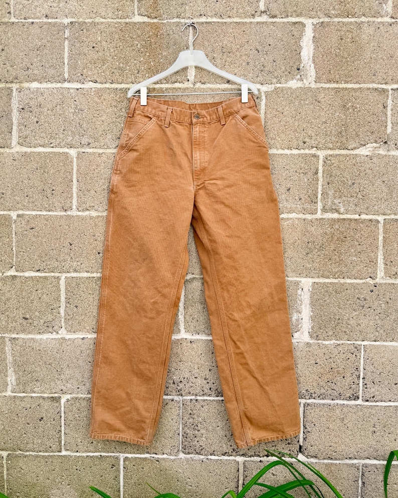 May include: A pair of tan-colored work pants hanging on a white hanger against a brick wall. The pants have a classic five-pocket design with reinforced stitching and a straight leg. The fabric appears to be a durable cotton blend.