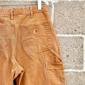 May include: Close-up of brown work pants with multiple pockets and a small leather patch with the Carhartt logo. The pants are hanging on a white hanger against a textured brick wall.