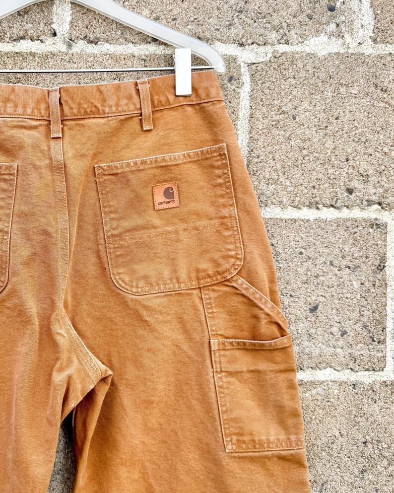 May include: Close-up of brown work pants with multiple pockets and a small leather patch with the Carhartt logo. The pants are hanging on a white hanger against a textured brick wall.