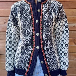 Könnte beinhalten: Eine dunkelblaue und weiße Strickjacke mit geometrischem Muster auf dem Körper und den Ärmeln. Die Knopfleiste ist mit einer dekorativen rot-grünen Stickerei verziert und verfügt über silberfarbene Metallknöpfe auf der Vorderseite.