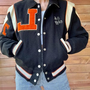 Peut inclure: Veste de baseball noire avec des rayures blanches et orange. La veste a un grand "L" orange sur le côté gauche et le nom "Cliff" brodé sur le côté droit.