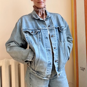 May include: Light blue denim jacket and matching jeans. The jacket has a classic collar, button-up front, and two chest pockets. The jeans are a straight-leg style. A patterned scarf is worn around the neck.
