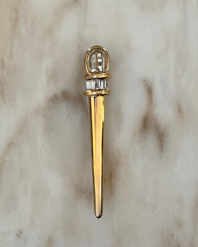 May include: A gold-colored letter opener with a decorative top and a pointed end. The top features a clear, rounded crystal design above a band of rectangular crystals. The opener is designed for opening envelopes and letters.
