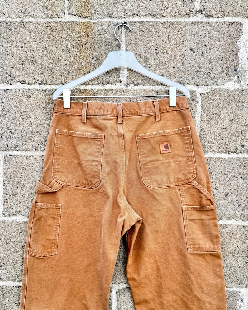 May include: Close-up of brown work pants with multiple pockets, hanging on a white hanger. The pants have a faded appearance and a small Carhartt logo patch on the back pocket. The pants are made of a durable material.