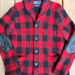Könnte beinhalten: Ein rot-schwarz karierter Cardigan mit Schalkragen und schwarzen Ellbogenpatches aus Leder. Der Pullover hat eine Knopfleiste vorne und zwei Vordertaschen. Das Polo by Ralph Lauren-Label ist sichtbar.