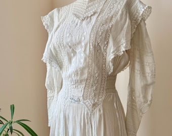 May include: A vintage white dress with intricate lace and embroidery details. The dress features a high neckline, long sleeves, and a flowing skirt. The bodice is adorned with delicate lace panels and floral embroidery. The dress is displayed on a mannequin.