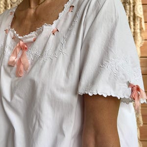 May include: A white cotton blouse featuring short sleeves with scalloped edges and pink ribbon accents threaded through eyelets. The neckline and sleeves are decorated with white floral embroidery, and the garment has a loose, relaxed fit.