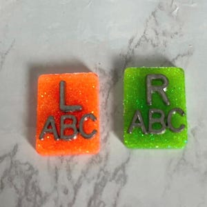 May include: Two small square tiles with glitter. One tile is orange with the letters "L ABC" and the other is green with the letters "R ABC".