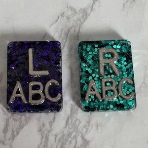 May include: Two square resin eye chart tiles with glitter embedded in the resin. The left tile is purple with the letter "L" and "ABC" printed in silver. The right tile is teal with the letter "R" and "ABC" printed in silver.