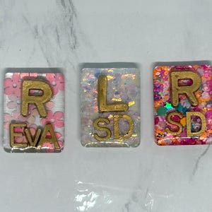 Può includere: Tre ciondoli rettangolari in resina con scritte dorate. Il primo ha le lettere "R" e "EVA" su uno sfondo floreale rosa. Il secondo ha "L" e "SD" su uno sfondo bianco con glitter. Il terzo ha "R" e "SD" su uno sfondo colorato.