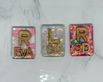 May include: Three rectangular resin pendants with gold lettering. The first has the letters "R" and "EVA" over a pink floral background. The second has "L" and "SD" over a white background with glitter. The third has "R" and "SD" over a colourful background.